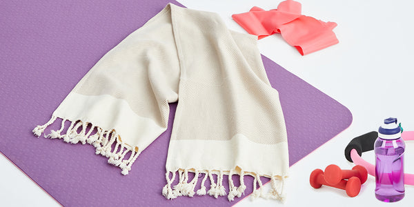 White towel with tassels on a purple mat with pink resistance bands and red dumbbells.